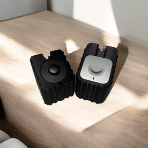May include: Two black and silver round knobbed devices with a ribbed texture. The devices are on a light wood surface.