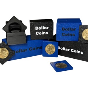 May include: A collection of coin display boxes in black and blue, some with gold dollar coins on display. The boxes are labelled "Dollar Coins" in white text. The arrangement includes various shapes and sizes.