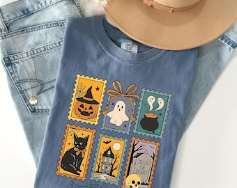 May include: A blue Comfort Colors t-shirt featuring a Halloween-themed stamp design. The stamps include a jack-o'-lantern, ghost, cauldron, black cat, haunted house, and skull with the words "Trick or Treat". The shirt is paired with light wash jeans and a tan hat.