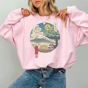May include: Pink sweatshirt featuring a Japanese-inspired graphic. The design includes a mountain, a full moon, and a tree with text in Japanese. The sweatshirt is a light pink color.