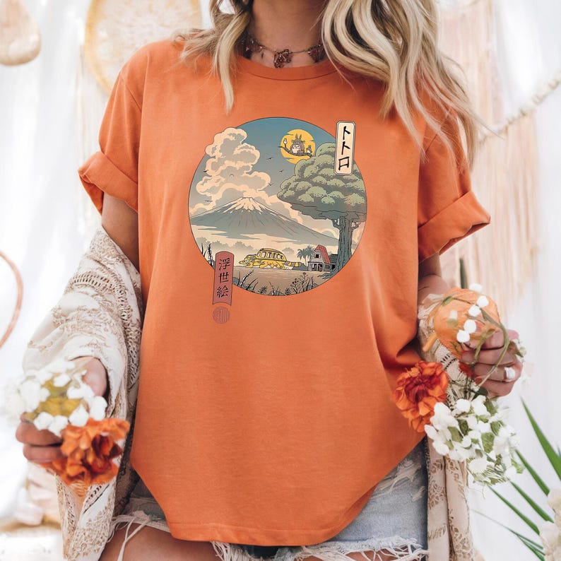 May include: Orange t-shirt featuring a circular graphic with a Japanese-inspired landscape. The design includes a mountain, tree, and characters, with Japanese text. The shirt has short sleeves and a relaxed fit.