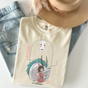 May include: Cream-colored t-shirt featuring a graphic from the animated film Spirited Away. The design includes a character with a white mask, a dragon, and a girl in a red outfit. The shirt is paired with blue jeans and a tan hat.