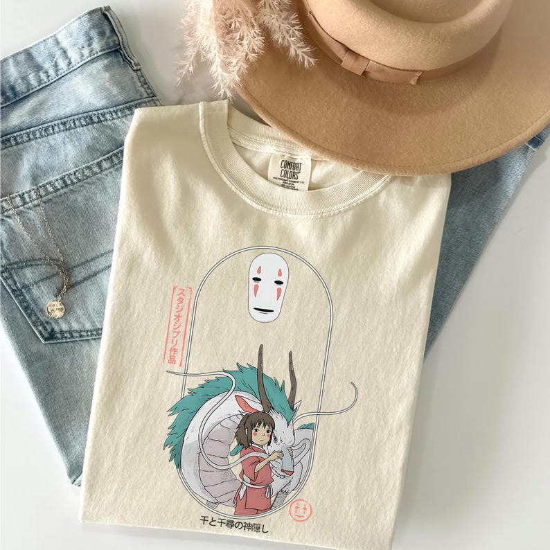 May include: Cream-colored t-shirt featuring a graphic from the animated film Spirited Away. The design includes a character with a white mask, a dragon, and a girl in a red outfit. The shirt is paired with blue jeans and a tan hat.