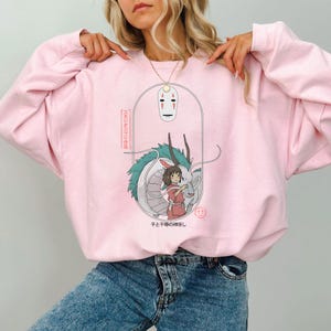 May include: Light pink sweatshirt featuring a graphic from the animated film, Spirited Away. The design includes a white mask, a dragon, and a girl in a red dress. Japanese text is also included in the design.