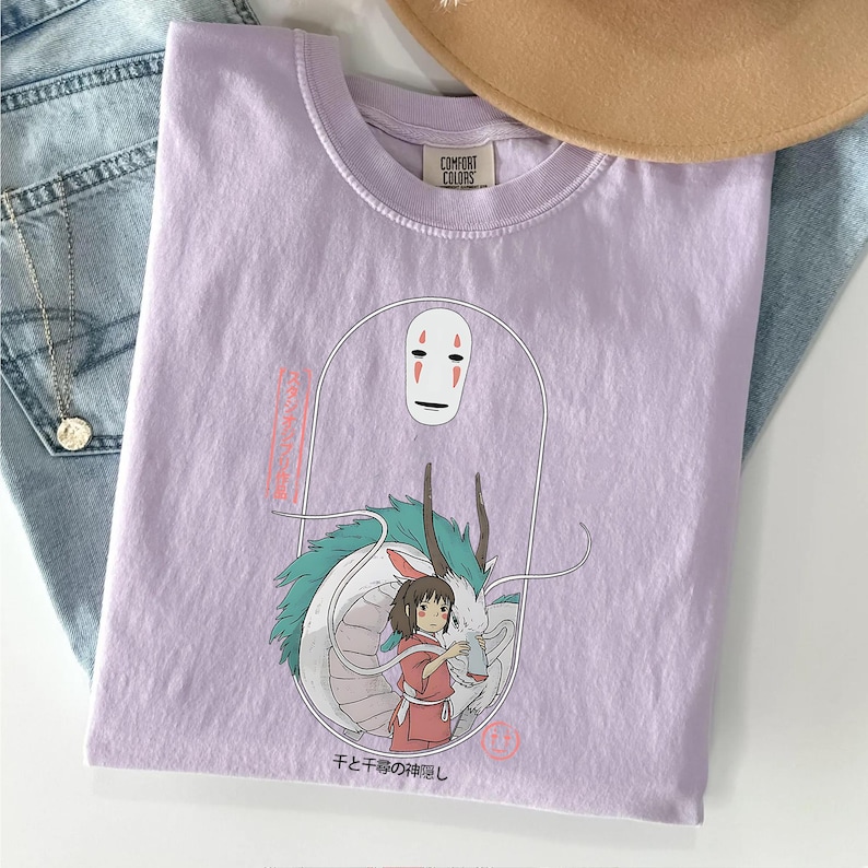 May include: A lilac Comfort Colors t-shirt featuring a graphic from the animated film Spirited Away. The design includes the character No-Face, a girl, and a dragon. Japanese text is also present.