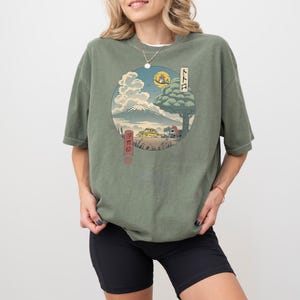 May include: Olive green t-shirt featuring a circular graphic of a Japanese landscape with a mountain, tree, and buildings. The graphic includes Japanese text and a full moon. The shirt is worn by a person.