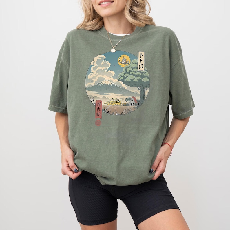 May include: Olive green t-shirt featuring a circular graphic of a Japanese landscape with a mountain, tree, and buildings. The graphic includes Japanese text and a full moon. The shirt is worn by a person.