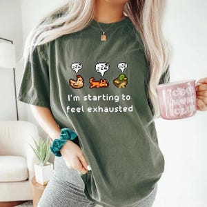 May include: Dark green t-shirt with a pixelated design of a chicken, cat, and duck with "zzz" speech bubbles above them. The text "I'm starting to feel exhausted" is below the images. A pink mug with text is held in the hand.
