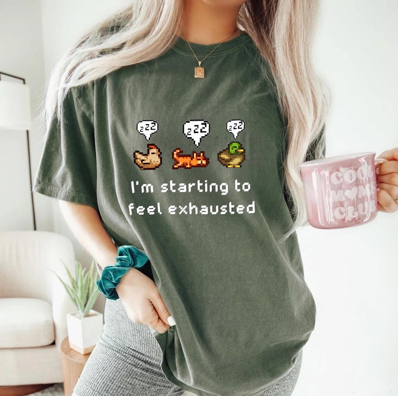 May include: Dark green t-shirt with a pixelated design of a chicken, cat, and duck with "zzz" speech bubbles above them. The text "I'm starting to feel exhausted" is below the images. A pink mug with text is held in the hand.