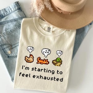 May include: Cream-colored t-shirt with pixelated chicken, cat, and duck graphics, each with a "zzz" speech bubble. The text "I'm starting to feel exhausted" is printed below the graphics. A tan hat and blue jeans are in the background.