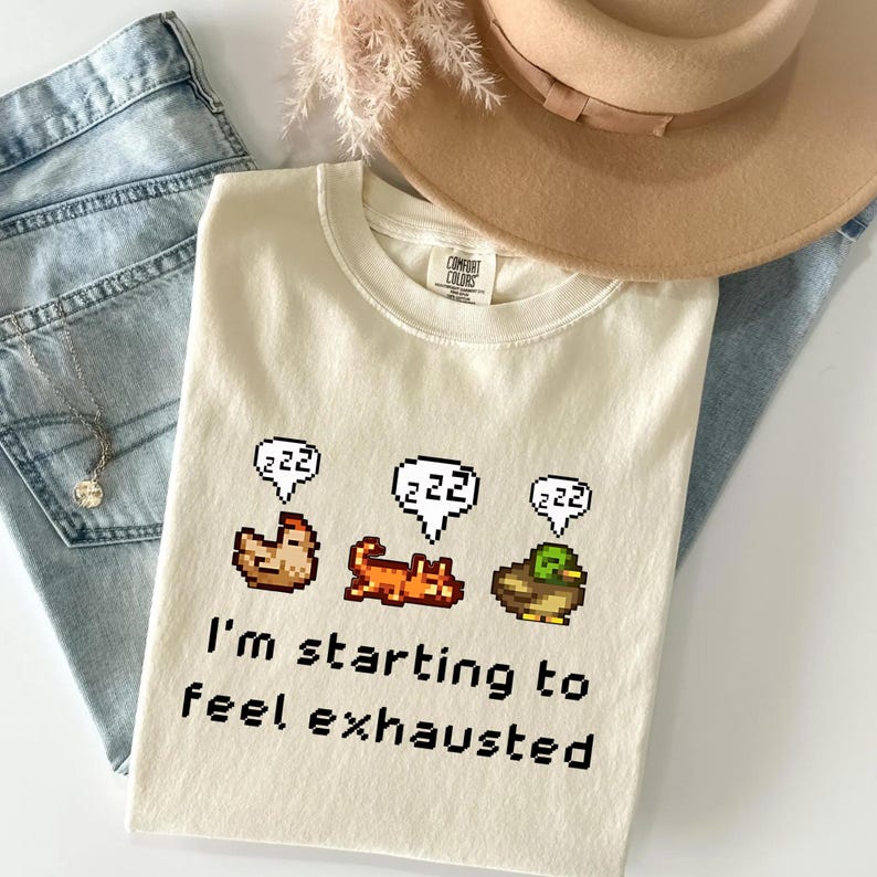 May include: Cream-colored t-shirt with pixelated chicken, cat, and duck graphics, each with a "zzz" speech bubble. The text "I'm starting to feel exhausted" is printed below the graphics. A tan hat and blue jeans are in the background.