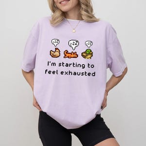 May include: A light purple t-shirt with a pixelated graphic of a chicken, cat, and duck, each with a "zzz" speech bubble. The text "I'm starting to feel exhausted" is below the graphic.