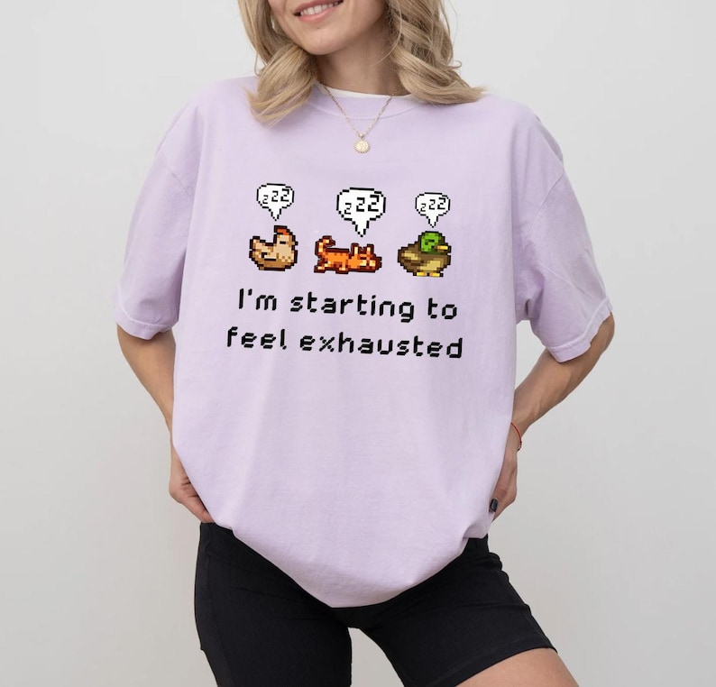 May include: A light purple t-shirt with a pixelated graphic of a chicken, cat, and duck, each with a "zzz" speech bubble. The text "I'm starting to feel exhausted" is below the graphic.