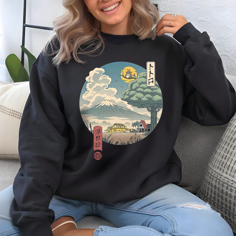 May include: Dark gray sweatshirt featuring a circular graphic with Japanese art elements. The design includes a mountain, clouds, a tree, and a yellow vehicle. Japanese text is also present. The sweatshirt is a comfortable, casual style.