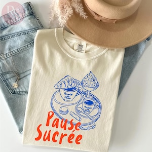 May include: Cream-colored t-shirt with a blue illustration of coffee, a croissant, and a sundae on a tray. The words "Pause Sucrée" are in orange. The shirt is laid on top of blue jeans and next to a tan hat.