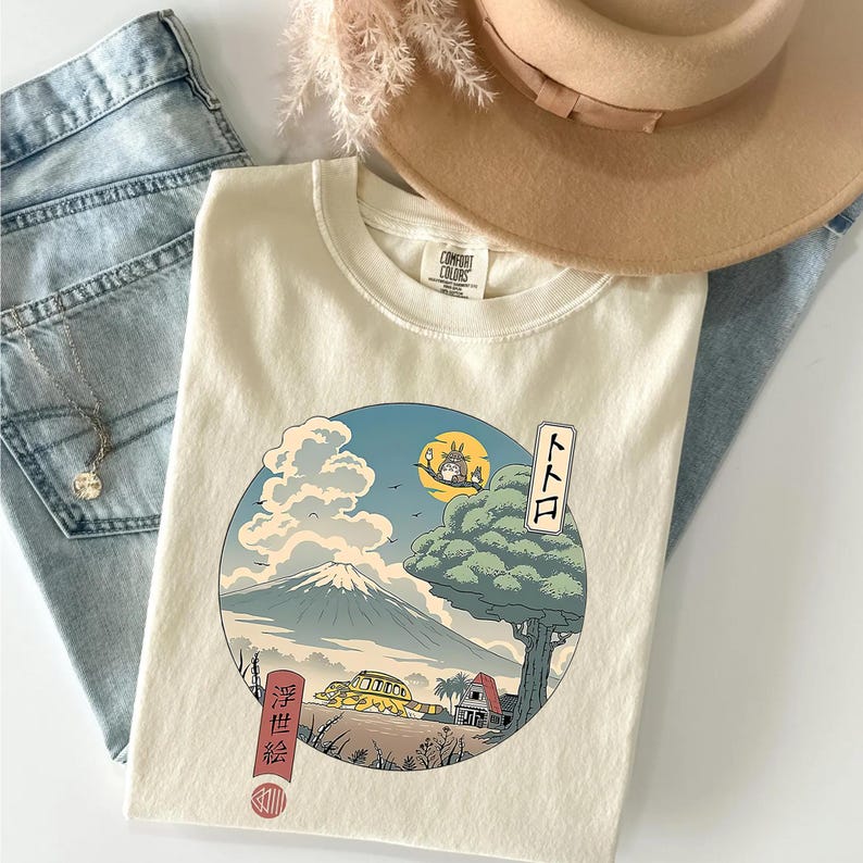 May include: Cream-colored t-shirt featuring a Japanese-inspired graphic with a mountain, tree, and a cartoon character. The shirt is folded with a hat and jeans in the background. The shirt has the text "トトロ" and Japanese characters.