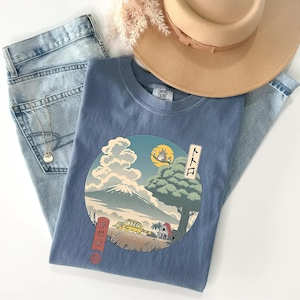 May include: A blue Comfort Colors t-shirt featuring a circular graphic with a Japanese-inspired landscape, including a mountain, tree, and a yellow vehicle. The shirt is paired with light-wash jeans and a tan hat.