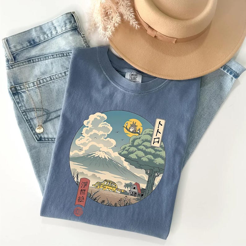 May include: A blue Comfort Colors t-shirt featuring a circular graphic with a Japanese-inspired landscape, including a mountain, tree, and a yellow vehicle. The shirt is paired with light-wash jeans and a tan hat.
