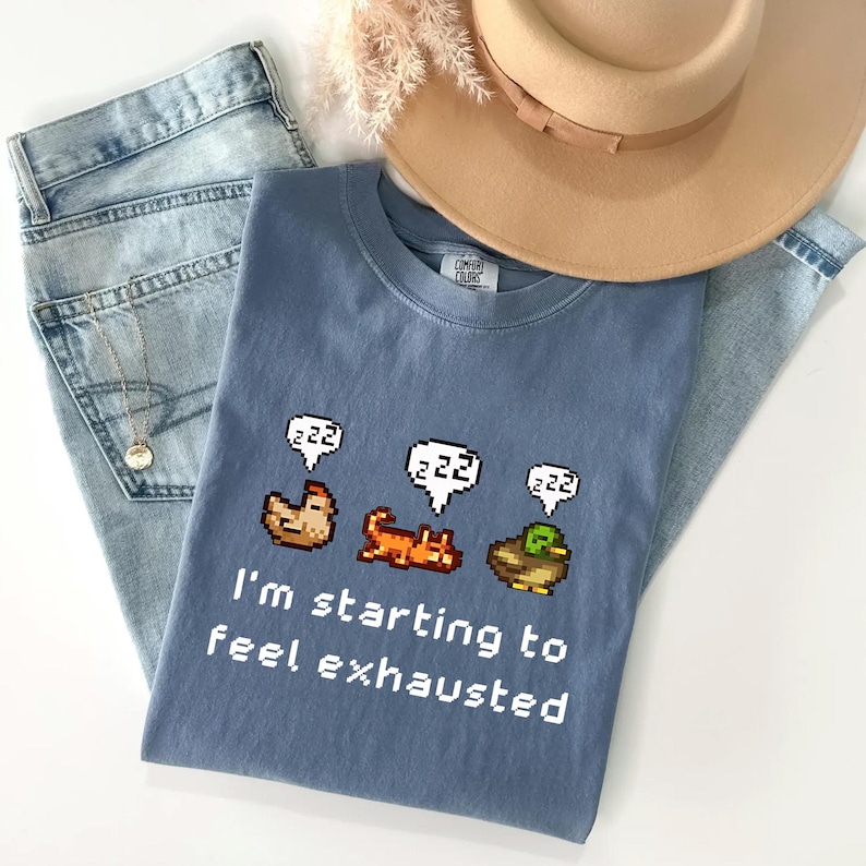 May include: A dusty blue t-shirt with a graphic of a chicken, cat, and duck, each with a "zzz" speech bubble. The text "I'm starting to feel exhausted" is below the graphics. The shirt is laid on top of light blue jeans and a tan hat.