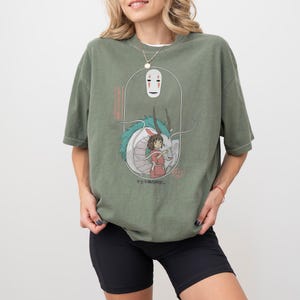 May include: Olive green t-shirt featuring a graphic from the animated film Spirited Away. The design includes the character No-Face, a dragon, and a girl in a red dress. Japanese text is also visible. The shirt has short sleeves.
