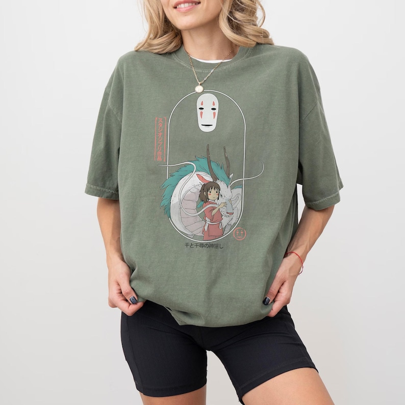 May include: Olive green t-shirt featuring a graphic from the animated film Spirited Away. The design includes the character No-Face, a dragon, and a girl in a red dress. Japanese text is also visible. The shirt has short sleeves.