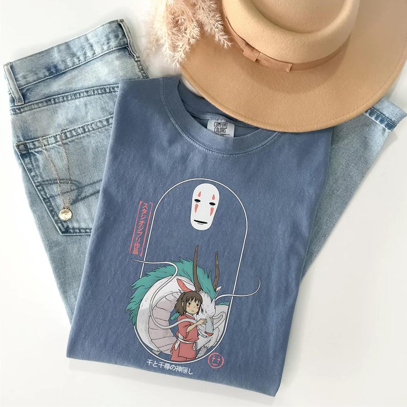 May include: A blue Comfort Colors t-shirt featuring a graphic from the movie Spirited Away. The graphic includes a white, faceless character, a girl in a red dress, and a white dragon. The shirt is paired with light wash jeans and a tan hat.