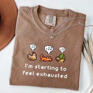 May include: A brown Comfort Colors t-shirt with the text "I'm starting to feel exhausted" and pixelated animal graphics. A silver necklace with a pendant is on the shirt. The shirt is a size small.