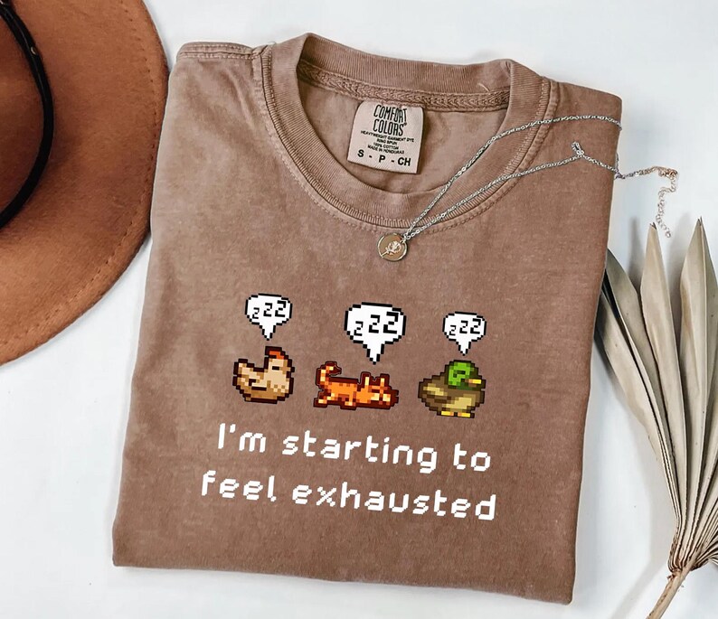 May include: A brown Comfort Colors t-shirt with the text "I'm starting to feel exhausted" and pixelated animal graphics. A silver necklace with a pendant is on the shirt. The shirt is a size small.