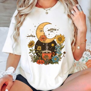 May include: White t-shirt with a graphic of a crescent moon with a face, a steaming cup of coffee, and flowers. The moon is surrounded by stars and clouds.