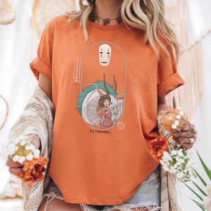 May include: Orange t-shirt featuring a cartoon illustration from the movie Spirited Away. The design includes the character No-Face, a girl in a pink dress, and a dragon. Japanese text is also present.