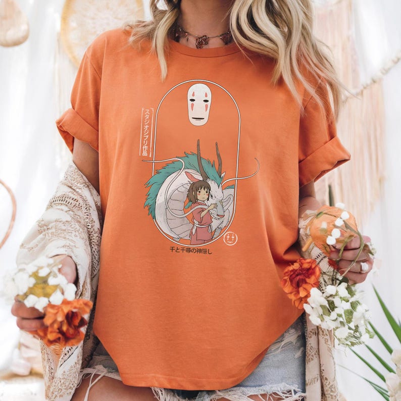 May include: Orange t-shirt featuring a cartoon illustration from the movie Spirited Away. The design includes the character No-Face, a girl in a pink dress, and a dragon. Japanese text is also present.