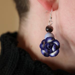 May include: A pair of earrings featuring a purple and white flower charm, a light blue bead, and a dark brown bead. The earrings are silver and have a hook closure.