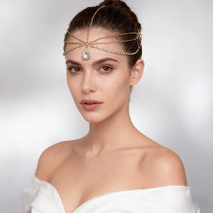 May include: A gold-toned headpiece with a central teardrop-shaped crystal and smaller crystals along the forehead. The headpiece is worn on the head and is part of a bridal or formal wear ensemble.