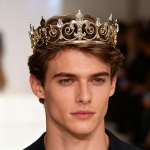 May include: A gold crown with elaborate detailing, including scrollwork and clear gemstones. The crown has a fleur-de-lis design and is suitable for a costume or special event. The intricate design suggests a royal theme.