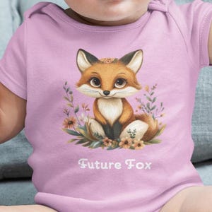 May include: A pink baby onesie with a cute cartoon fox wearing a flower crown. The fox is surrounded by flowers and greenery. The onesie has the text "Future Fox" printed on the front.