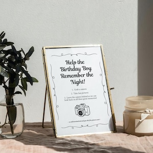 May include: A gold-framed sign with the text "Help the Birthday Boy Remember the Night!" and instructions to take pictures with a camera. The sign is on a table with a vase of greenery and a candle.