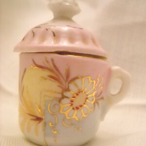 May include: A small, pink and white porcelain creamer with a lid. The creamer is decorated with gold floral designs and a delicate floral pattern.