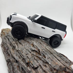 May include: A white and black toy SUV with black wheels is positioned on a piece of wood. The SUV has a detailed design, including a front grill, side steps, and a black roof. The wood has a rough, textured surface.
