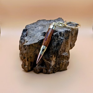 May include: A handcrafted wooden pen with a gold-colored metal tip and accents, attached to a gold keychain. The pen features a rich, dark brown wood grain and is displayed on a textured, gray stone.