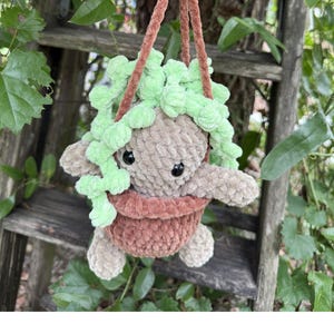 May include: A crocheted plush figure with a tan body and black eyes, nestled inside a brown yarn pot. The figure is adorned with a cascading light green yarn plant and is suspended by a brown yarn hanger, creating a soft, textured appearance.