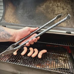 May include: A pair of long, dark grey metal barbecue tongs featuring a textured, twisted handle design and claw-shaped tips. The tongs are held over a metal grill grate containing four raw, light pink sausages arranged in a horizontal row.