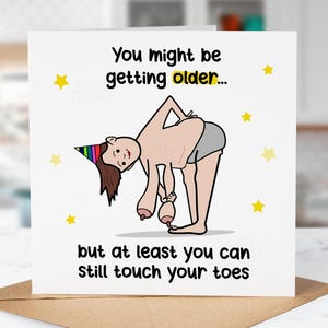 May include: A funny birthday card featuring a cartoon figure bending over, touching their toes. The card reads "You might be getting older... but at least you can still touch your toes". Yellow star decorations are also present.