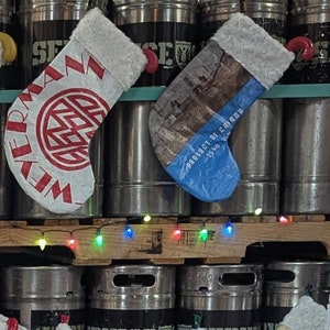 May include: A stack of stainless steel kegs decorated with Christmas stockings. The stockings are made from repurposed grain bags and feature various brewery logos and branding. The kegs are adorned with a string of Christmas lights.