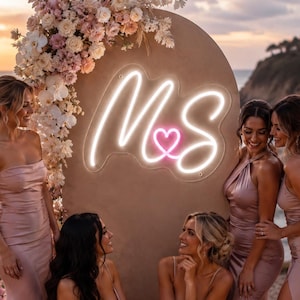 May include: A custom neon sign featuring the white glowing letters M and S connected by a pink heart symbol, mounted on a large, arched, matte tan backdrop decorated with a cascading arrangement of white and soft pink floral blossoms.