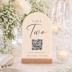 May include: An arched cream-colored table sign with a wooden base featuring the text TABLE Two, a QR code, the phrase Share your favorite moments, and the names Olivia & James with the date 09.14.2026 printed in black ink on the front surface.