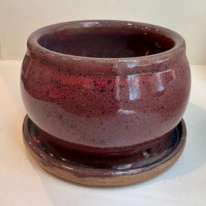 May include: A round ceramic planter with a glossy, speckled deep burgundy and dark red glaze. The pot features a wide, open top and sits securely on a matching circular saucer with an exposed, unglazed tan clay rim at the base.