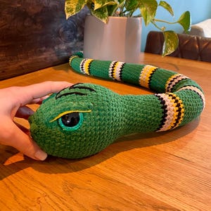 Op de afbeelding: Een groen gehaakt slangenknuffel met zwarte, gele en witte strepen. De kop van de slang is groen met zwarte markeringen en een gele oog. Het speelgoed is gemaakt van zacht garen en is ontworpen als kinderspeelgoed.