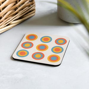 May include: Square coaster with a cream background and a pattern of colorful concentric circles in orange, yellow, and blue. The coaster has a black border and is on a light surface.