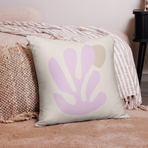 May include: A square decorative pillow with a light beige background. The pillow features a large, abstract design in soft pink and tan. The pillow is placed on a bed with a striped blanket and a woven throw pillow.