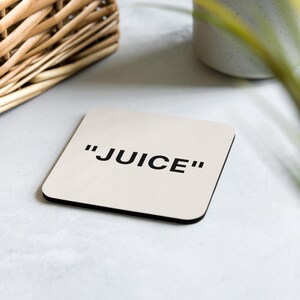 May include: Square off-white coaster with the word "JUICE" in bold black font. The coaster has a black border and is placed on a light gray surface. A woven basket and a ceramic pot are in the background.